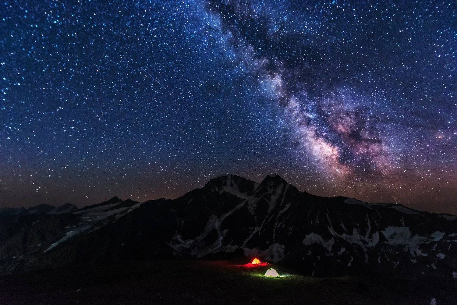 Milky Way over snow-dusted mountains with two illuminated camping tents on a plateau at night